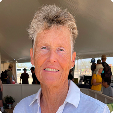 Smiling older woman with short, light hair wearing a white collared shirt under a tent with people in the background.