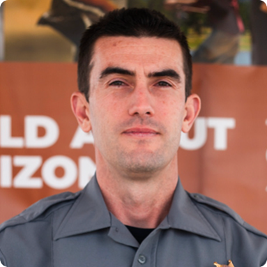 Man with short dark hair wearing a gray uniform shirt standing in front of a blurred brown background with partially visible white text.
