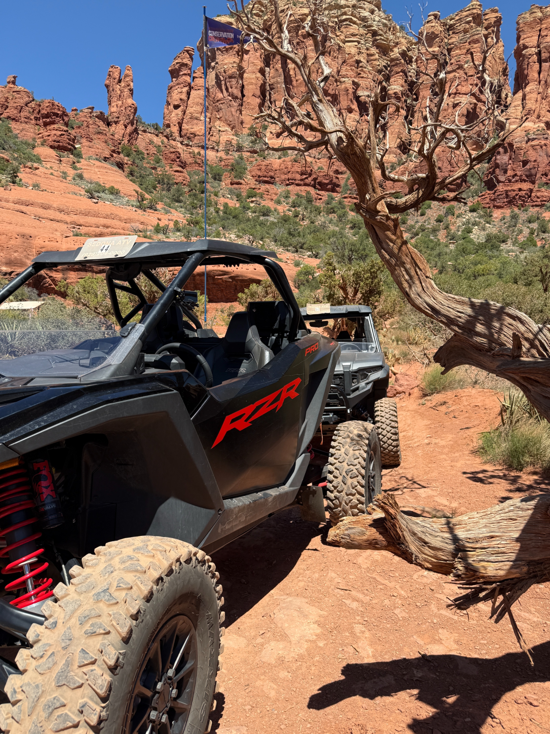 Two off-road RZR vehicles parked on a dusty red dirt trail with red rock formations and a twisted tree in the background under a clear blue sky.