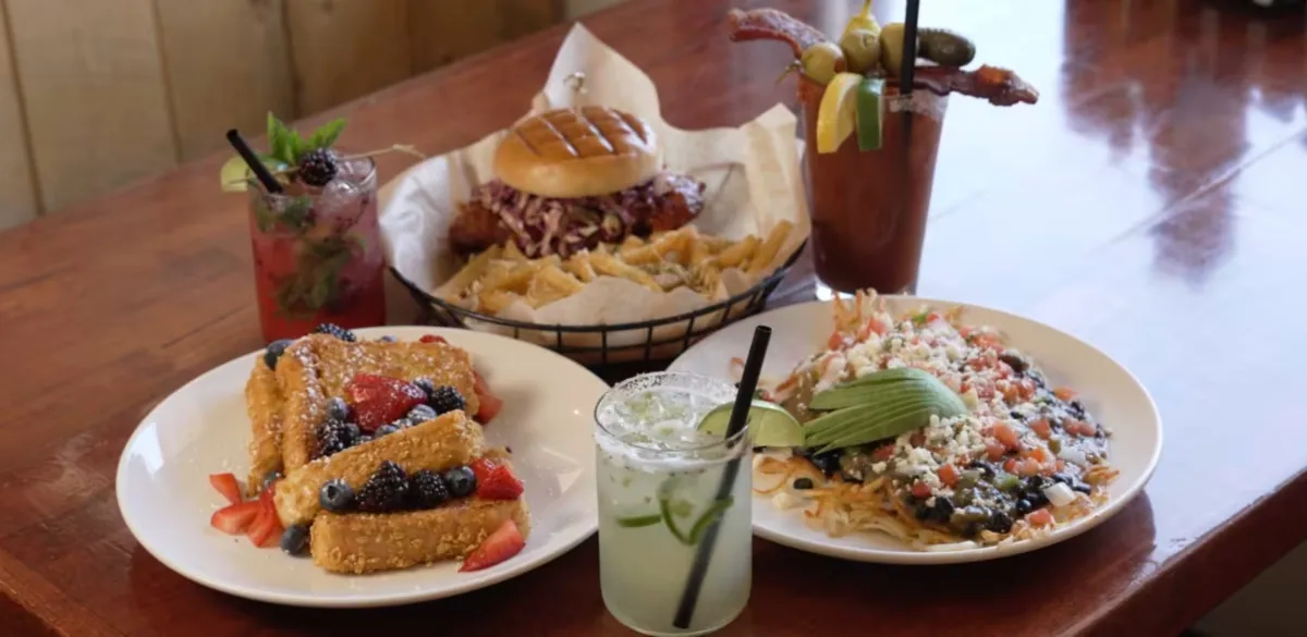 Outdoor patio seating area with fire pit, black chairs around it, picnic tables under a pergola, string lights, and mountains in the background at sunset.