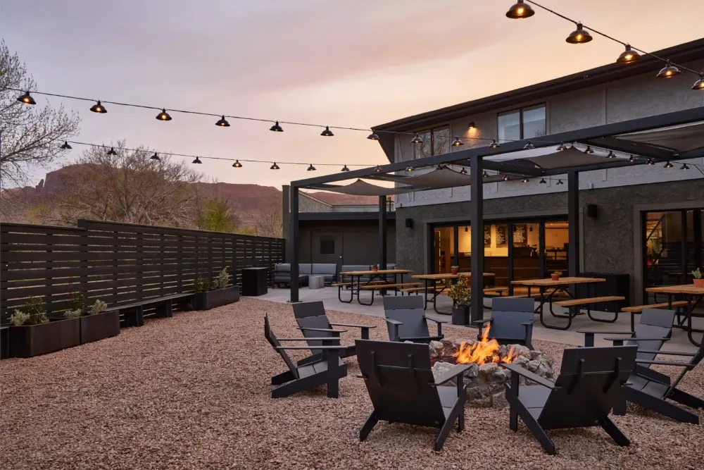 Outdoor patio seating area with fire pit, black chairs around it, picnic tables under a pergola, string lights, and mountains in the background at sunset.