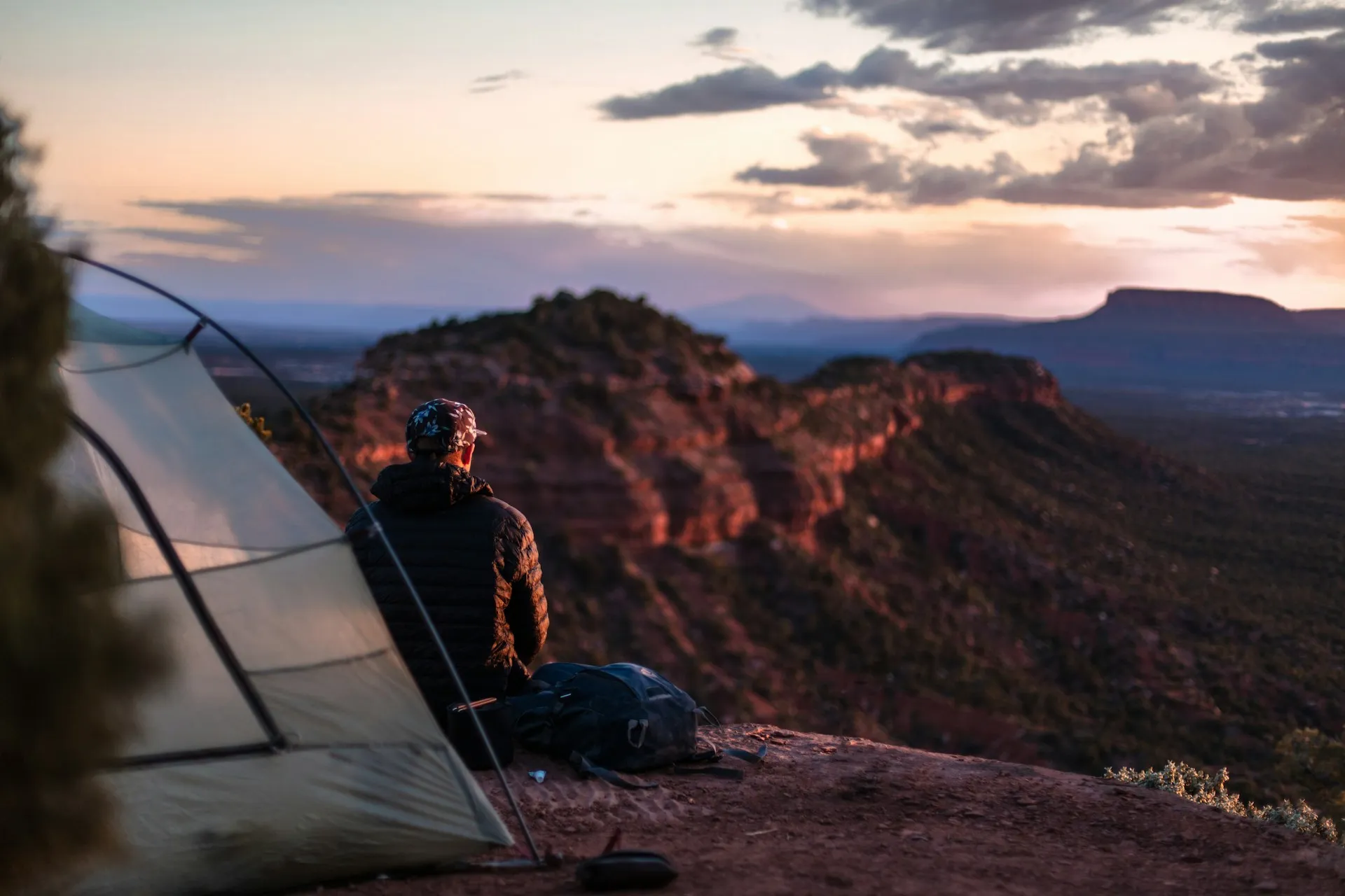 Person sitting next to a tent on a cliff, overlooking a vast landscape of rocky hills and a colorful sunset sky.
