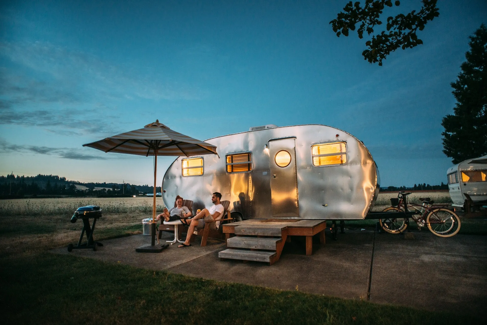 Two people sitting and relaxing on chairs under an umbrella outside a silver camper trailer at dusk.