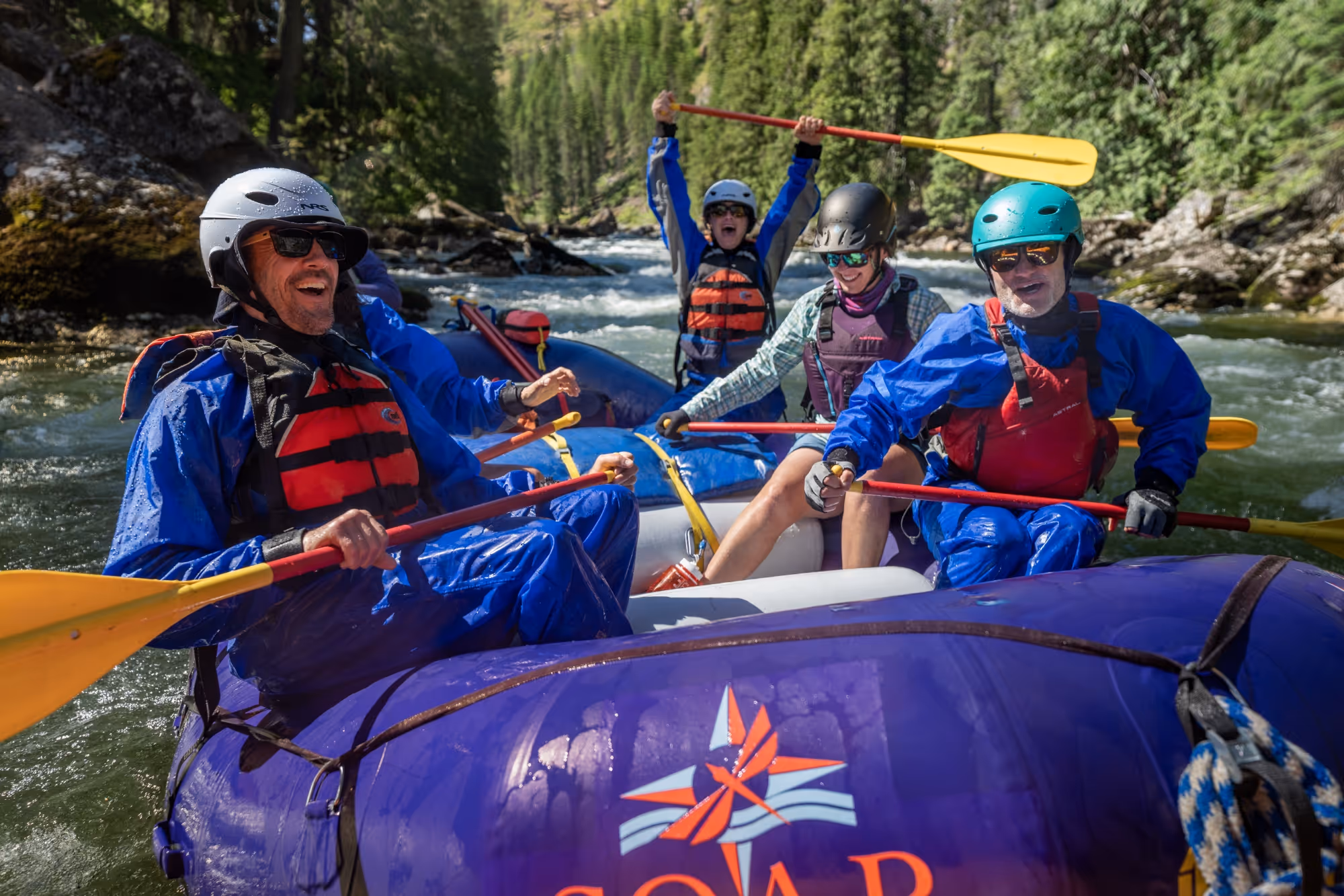A group of people in a red and black inflatable raft navigating whitewater rapids between rocky cliffs.