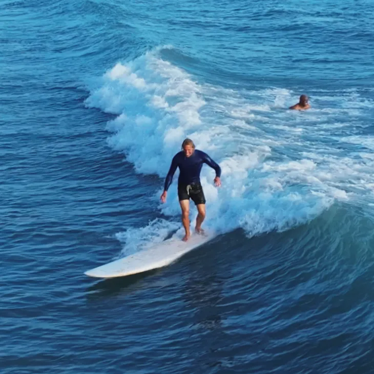 Surfer in a navy top riding a wave on a white surfboard in blue ocean water.