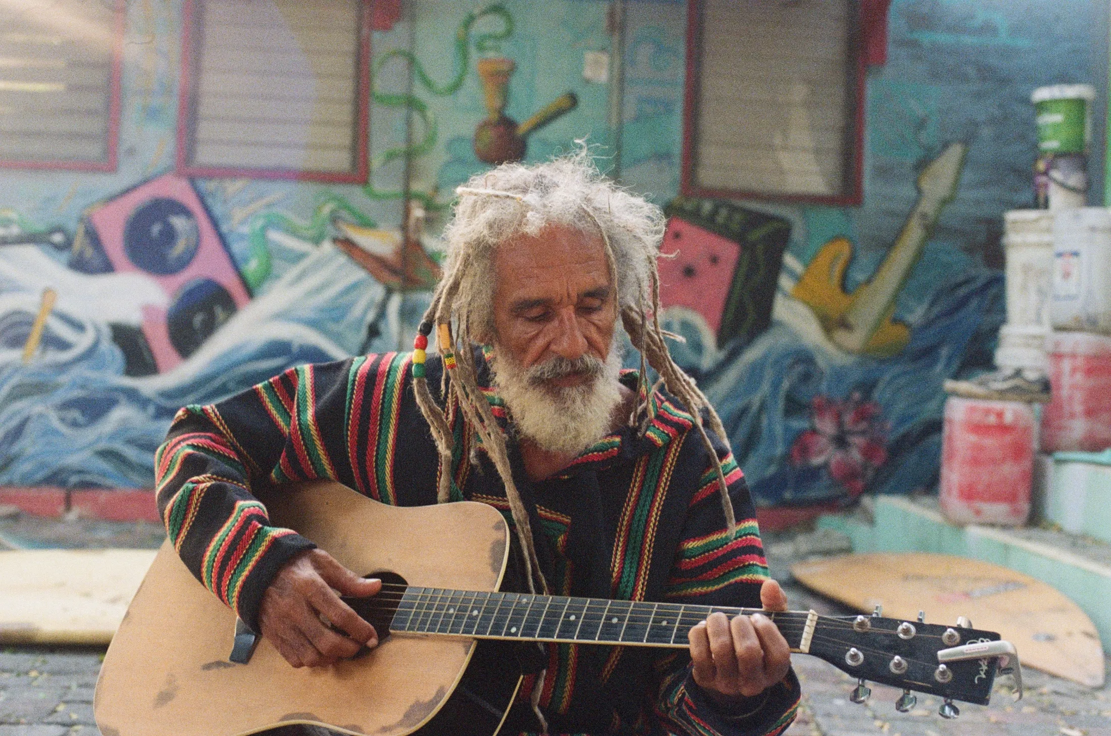 Elderly man with dreadlocks playing an acoustic guitar in front of a colorful mural.