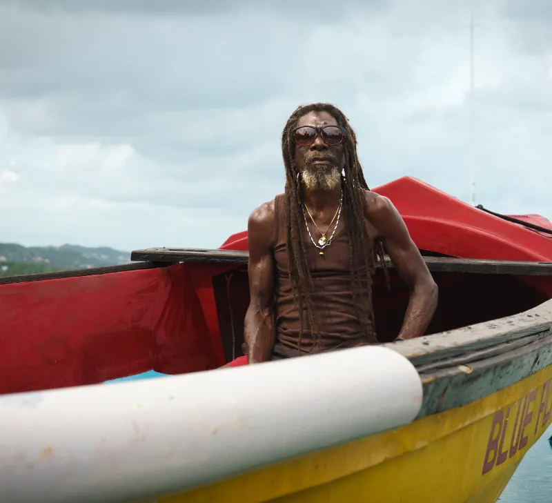 Man with long dreadlocks and sunglasses sitting in a yellow and red boat on water.