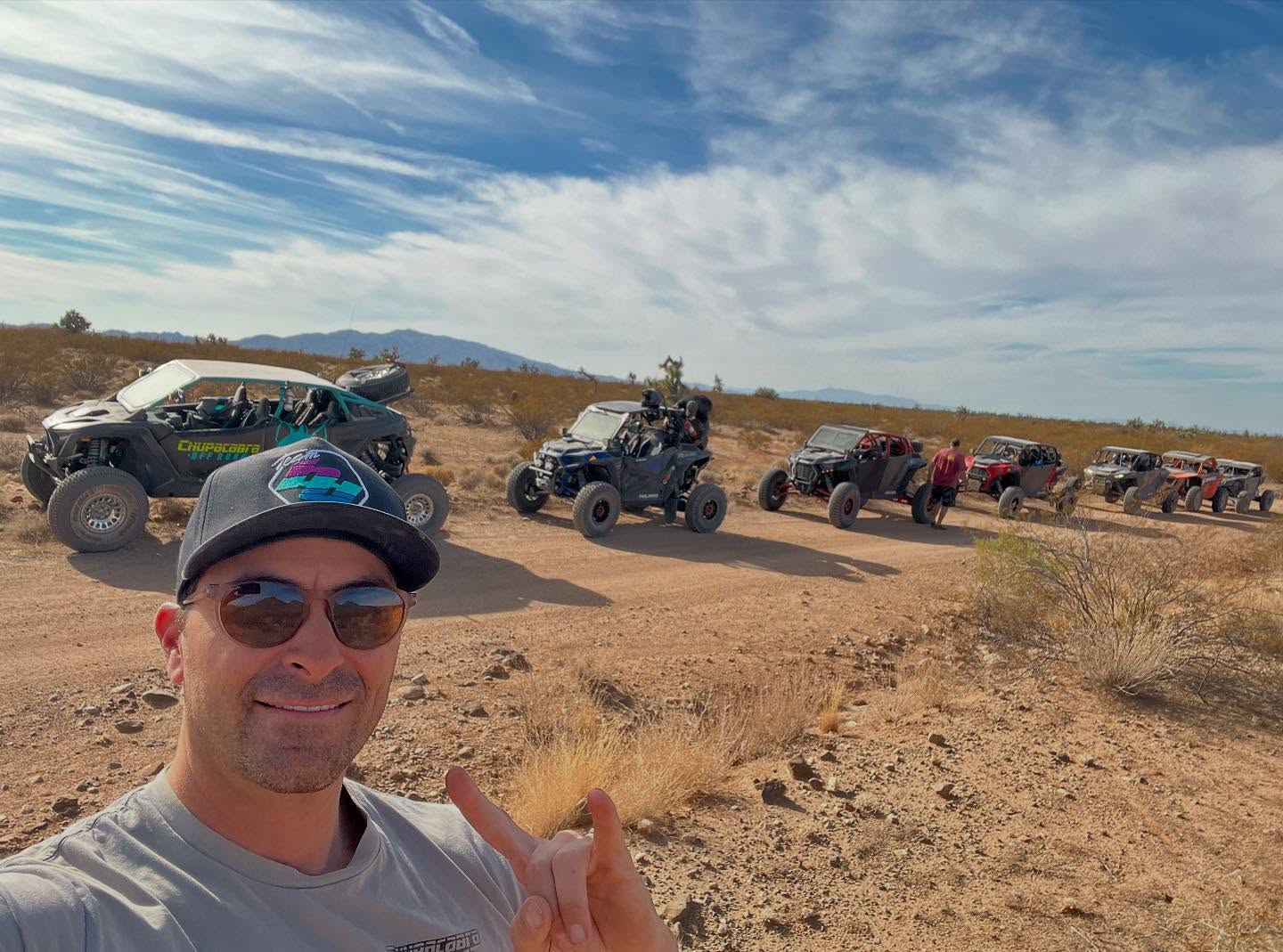 Man wearing sunglasses and a hat taking a selfie with six off-road vehicles lined up on a dirt trail in a desert landscape.